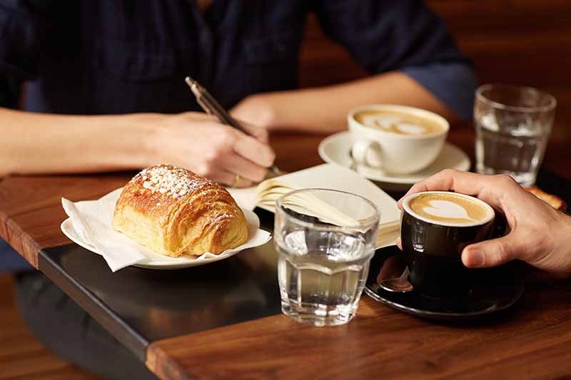 shot of two people sitting opposite each other in a coffee shop, with one person writing in a notebook and cups of coffee on the table